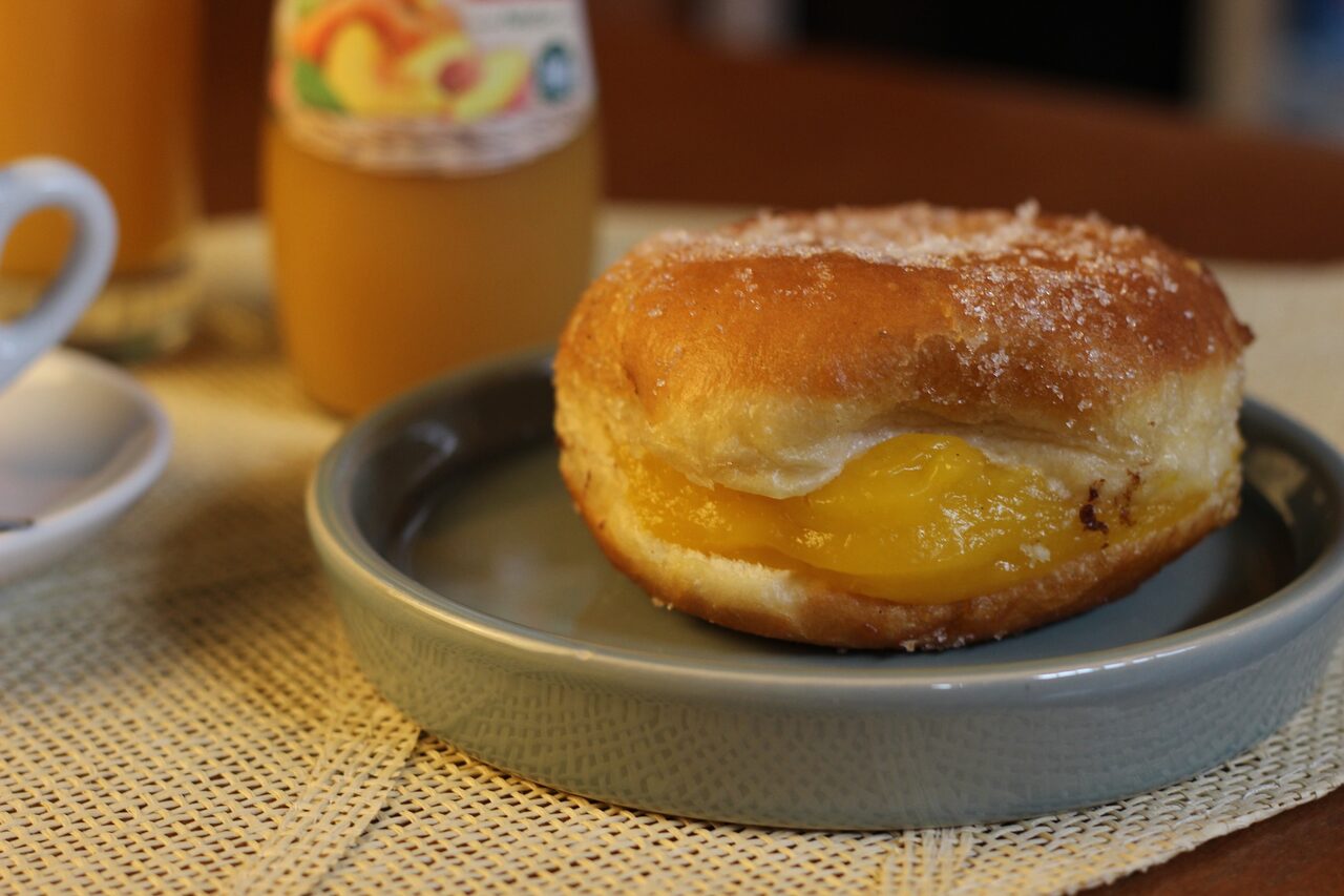 Uma bolinha de creme num prato cinzento, com o creme amarelo a sair pelo lado e um sumo desfocado atrás.