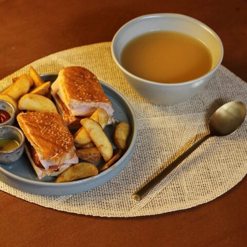 Cachorro em pão de sementes, cortado em dois, com batatas em gomos, molhos e uma tigela de sopa, sobre uma base de mesa em forma de folha.