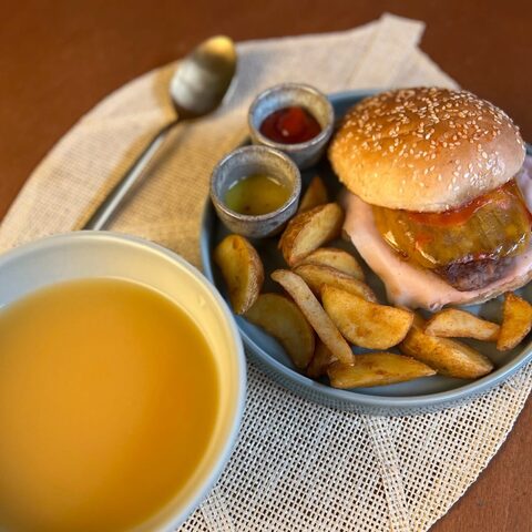 Hambúrguer em pão com sésamo, com batatas em gomos, molhos e uma tigela de sopa, sobre uma base de mesa entrançada.