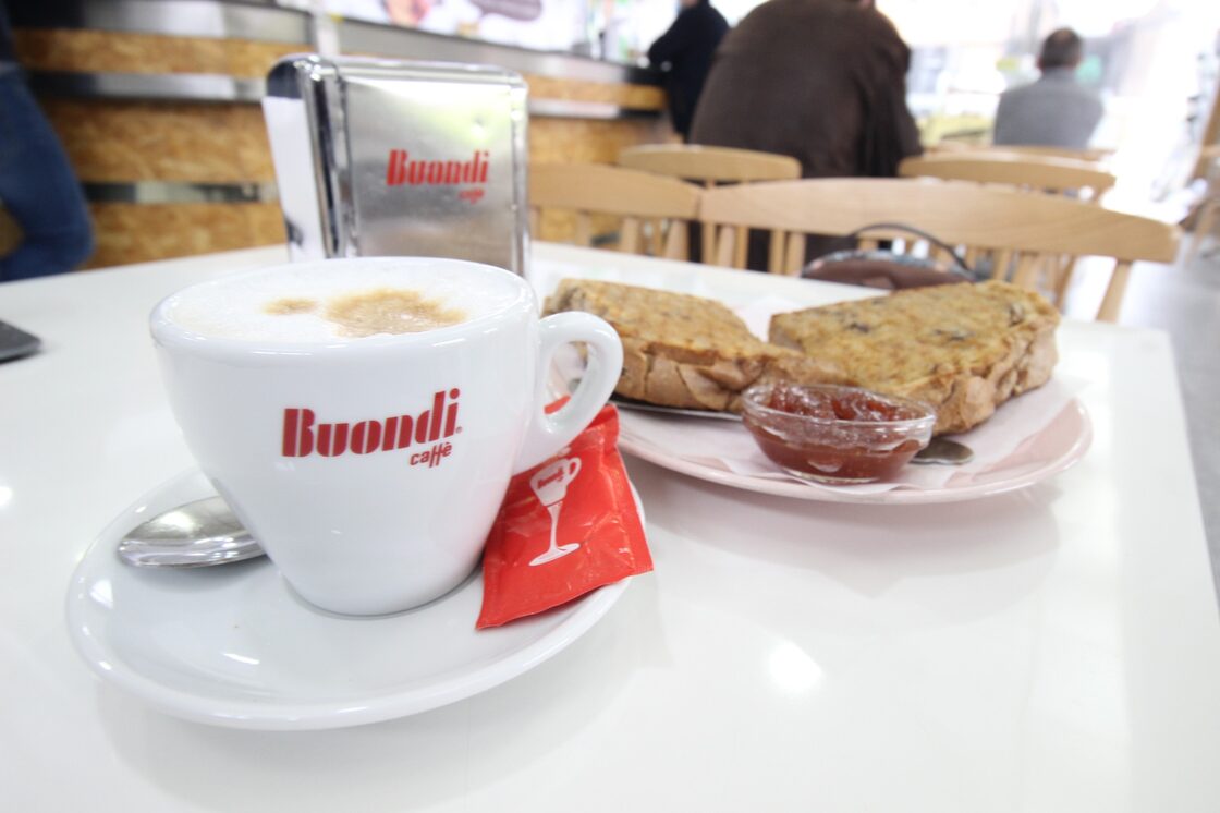 Meia de leite numa chávena Buondi e torrada com compota, numa mesa branca do Café Bocage, com a sala e as cadeiras de madeira atrás.