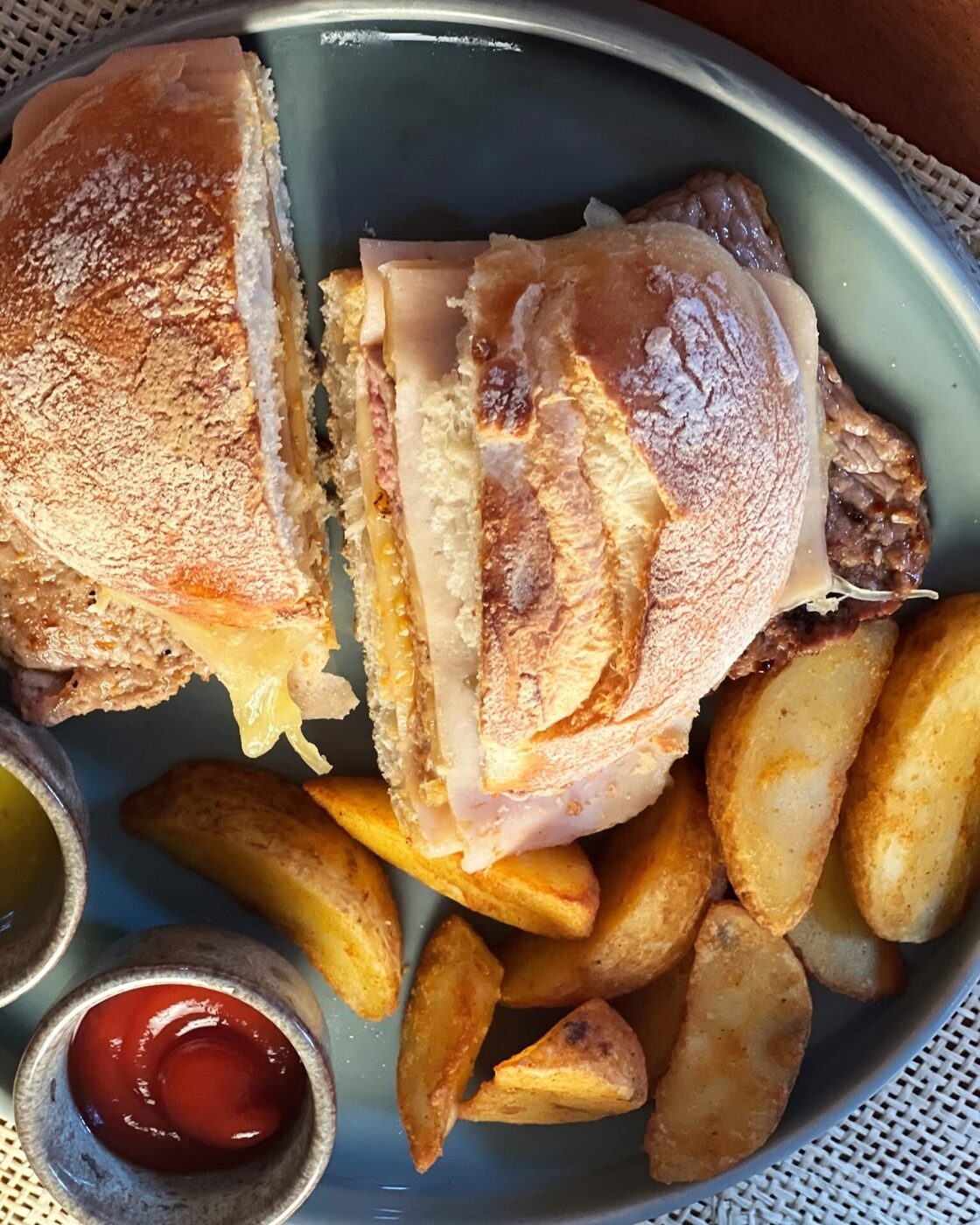 Prego no pão estaladiço, cortado ao meio, com batatas em gomos e duas taças de molho, num prato cinzento.