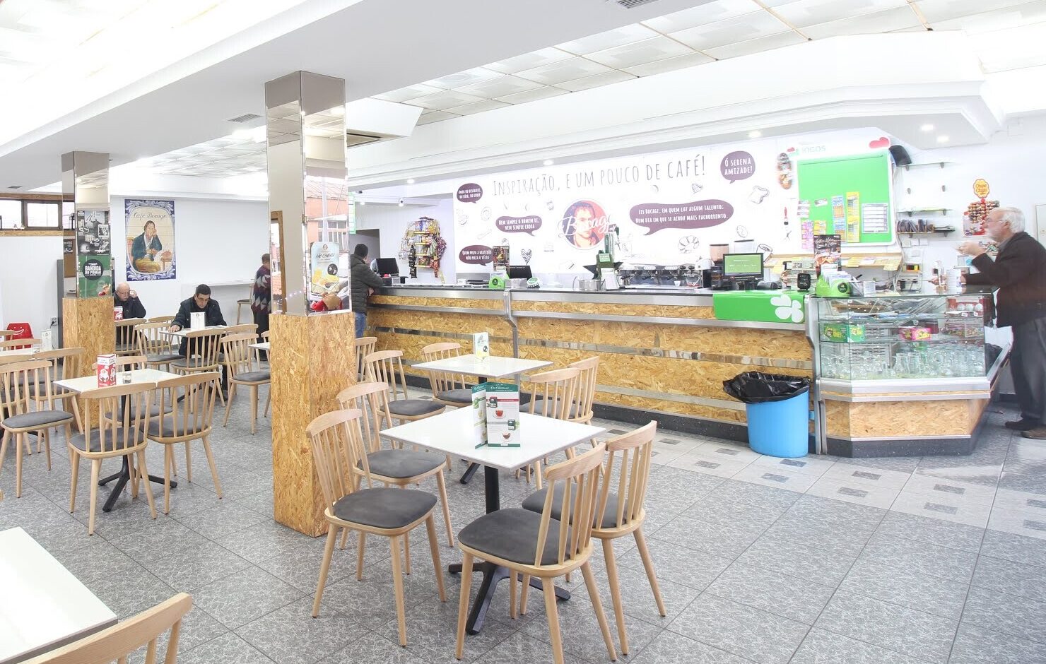 A sala do Café Bocage, clara e ampla, com cadeiras de madeira, o balcão comprido e, ao fundo, a parede com o retrato de Bocage e a frase «Inspiração, e um pouco de café!».