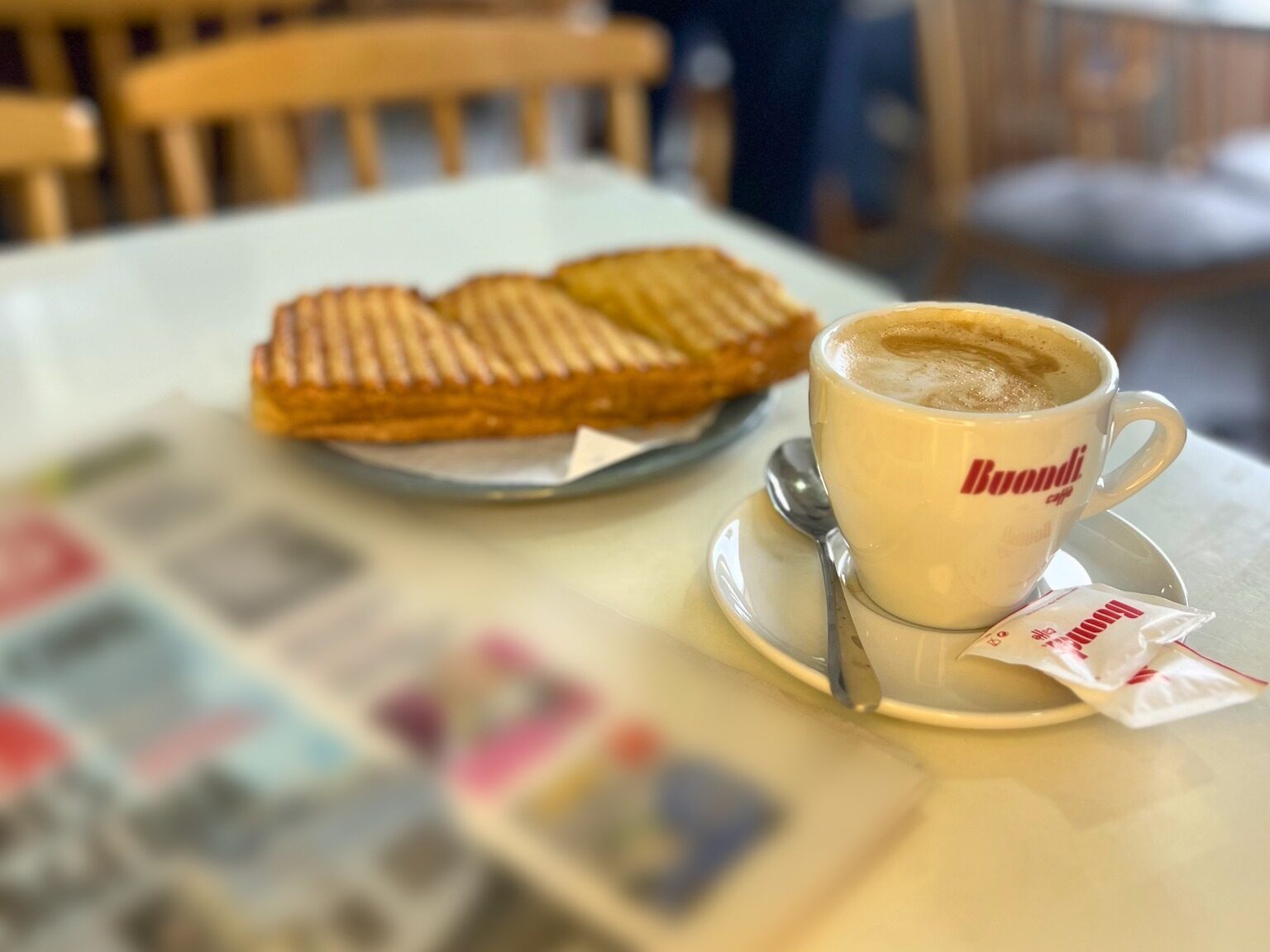 Meia de leite numa chávena Buondi e uma torrada num prato, numa mesa à luz da janela do Café Bocage, com cadeiras de madeira e clientes desfocados ao fundo.